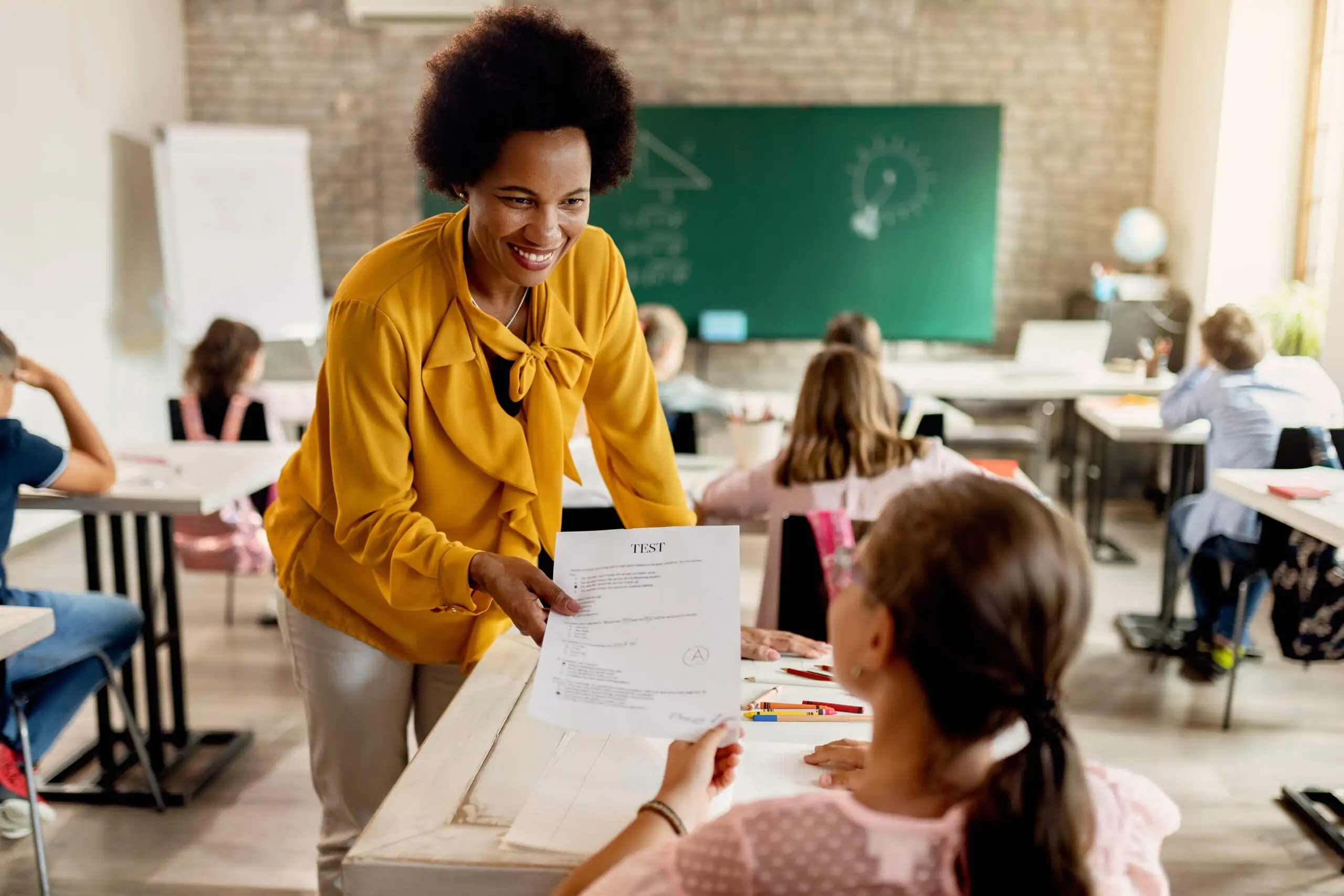 Happy African American teacher giving test results to elementary student in the classroom.